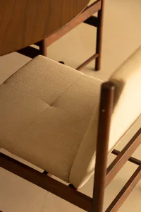 Close-up of a mid-century modern walnut dining chair featuring a beige textured fabric seat and backrest, with a curved walnut dining table visible in the background on a light-colored floor.