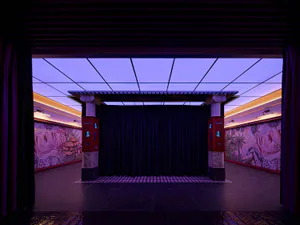 View from the audience into a contemporary theater stage featuring a black velvet curtain, two red-and-marble classical columns, large-scale surreal murals of animals and nature, and dramatic purple ceiling lighting.