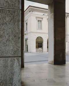 View through a textured stone archway toward a white neoclassical building with arched windows and decorative ironwork on a paved plaza.