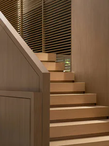 Modern minimalist wooden staircase with light oak treads and risers, featuring a solid oak handrail and a background wall of horizontal wooden slats allowing soft light to filter through.