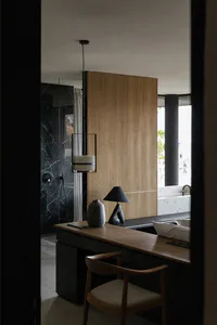 Modern bathroom interior viewed through a doorway, featuring a wooden vanity desk with a sculptural black lamp and ceramic vase, a wooden chair, a large warm wood panel wall, a black marble shower area, and a white terrazzo bathtub by a window.