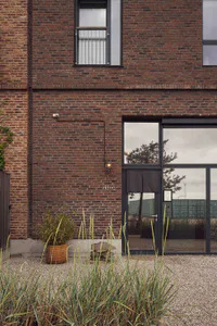 Exterior view of a red brick building with large modern glass doors, a wall-mounted light fixture, potted plants, and tall ornamental grasses in the foreground.