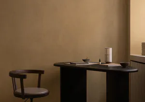 Minimalist home office corner with a dark walnut curved desk, wooden swivel chair with leather seat, ceramic bowls, table lamp, and books against a textured beige wall.