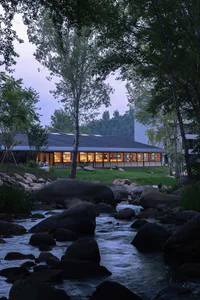 現代混凝土度假建築在黃昏時分，室內燈光透亮，旁邊有潺潺溪流與巨大岩石，四周被樹木環繞。