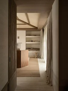 View down a narrow minimalist hallway featuring light oak flooring and walls, built-in white floating shelves, a small wooden stool, sheer linen curtains, and a woven ceiling treatment.
