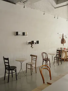 Bright minimalist cafe interior featuring a collection of wooden dining chairs including Thonet bentwood styles, small round tables, white tiled wainscoting, and textured plaster walls with floating shelves.