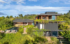 Exterior view of a modern tropical luxury villa with dark wooden and concrete facade, extensive green walls, infinity pool, and wooden decks nestled in a dense jungle landscape under a bright blue sky.