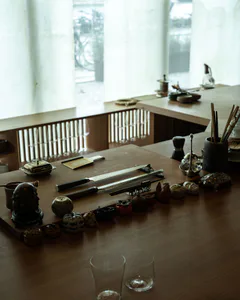 日式極簡風格的木製餐桌，擺放著陶瓷人偶、金屬雕像、武士刀與玻璃器皿，背景為大片窗戶。