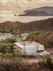 Modern white concrete villa with rooftop terrace and infinity pool overlooking the Pacific Ocean and forested cliffs at sunset.
