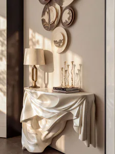 Modern sculptural console table with draped ivory finish, brass table lamp, and golden candlesticks against a neutral wall with decorative plates.