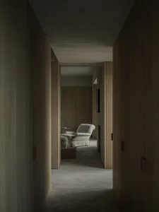 Long narrow hallway with vertical oak wood paneling on walls and ceiling, leading to a light-colored upholstered armchair and ottoman in a sunlit alcove with a woven basket on the concrete floor.