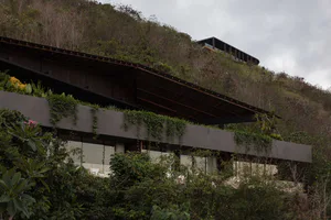 Modern concrete hillside villa with extensive green roof and living walls, nestled into a tropical forested slope under overcast skies.
