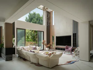 Modern double-height living room featuring a large curved white sectional sofa, marble floors, high ceilings with skylight, and large windows overlooking green trees.