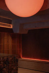 Interior view of a dimly lit speakeasy-style bar with dark red terrazzo walls, warm wood cabinetry, a stone bar counter, and a large glowing red spherical pendant light dominating the ceiling.