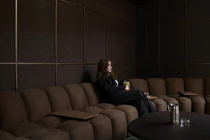 A woman with long brown hair wearing black sits on a brown modular sofa in a dark lounge, holding a glass of green matcha drink, surrounded by textured dark wall panels and a round coffee table with a metal pitcher.