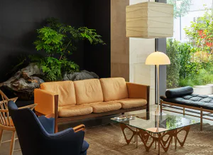 Modern living room featuring a tan leather three-seater sofa, dark blue armchair, sculptural walnut coffee table, large paper lantern pendant light, and indoor rock garden with lush greenery.