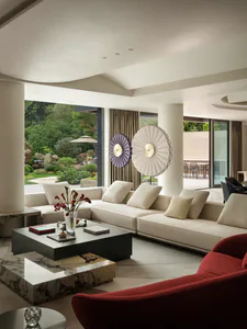 Modern luxury living room with a large L-shaped cream sectional sofa, black and marble coffee tables, a red velvet armchair in the foreground, two large circular pleated wall art pieces in purple and white, and large glass doors opening to a lush garden.