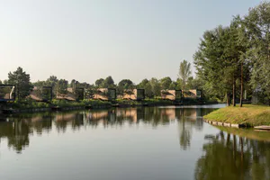 Row of modern wooden cabins with large glass windows situated along the edge of a calm pond surrounded by pine trees under a clear sky.