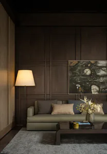 Modern living room featuring dark wood wall paneling, an olive-green sofa with neutral pillows, a tall floor lamp, abstract painting, and a dark wood coffee table on a grey rug.