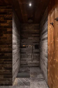 Narrow modern spa shower room with dark stacked slate stone tiles on walls and floor, warm vertical cedar wood paneling on the right, a large round rain shower head, handheld wand, built-in niche, and three black product bottles mounted on the back wall.