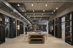 Modern industrial-style clothing boutique interior with raw concrete walls, black metal shelving, central wooden display table, and track lighting.