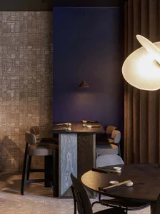 Modern Japanese-inspired restaurant interior with a high wooden dining table, upholstered bar stools, dark blue accent wall, patterned geometric tile wall, and a large sculptural pendant lamp in the foreground.