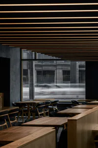 Modern minimalist restaurant interior featuring light oak wooden tables and benches, black upholstered chairs, dark concrete walls, and dramatic parallel linear LED lights on a black ceiling, with large windows showing motion-blurred city traffic outside.