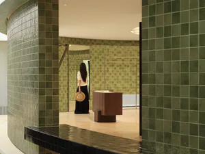 Woman standing in a modern boutique with curved walls covered in glossy green ceramic tiles and a minimalist walnut display counter.