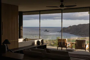 Modern open-plan living room in a cliffside villa featuring a terrazzo soaking tub, wooden lounge chairs on the terrace, a black ceiling fan, and dramatic ocean and mountain views through floor-to-ceiling glass walls.