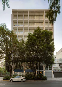多層混凝土公寓大樓正面視圖，帶有陽台、地面層大型拱形窗戶，前方有茂密綠樹，街道上停著一輛白色汽車。