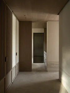 A long, narrow minimalist hallway featuring vertical wood slat doors, light oak cabinetry, travertine stone flooring and walls, and a dark-framed ribbed glass door at the end.