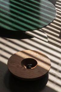 A round green marble coffee table with black base positioned above a circular oak wood tray containing a black truffle, both illuminated by strong sunlight creating striped shadows on a light wooden floor.