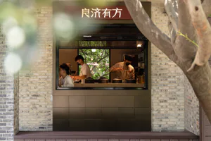 現代餐廳開放式廚房外觀，兩位廚師在深色磁磚吧台後準備餐點，以淺色磚牆為框，前景有樹木。