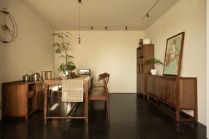 A minimalist dining room with dark walnut wooden furniture in Ming dynasty style, a long dining table with woven runner, wooden chairs, sideboard, tall bookshelf, potted plants, and a large framed painting of Guanyin on a beige wall with dark floor.