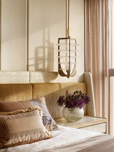 Close-up of a luxurious bedroom corner featuring a mustard-yellow velvet upholstered headboard, layered neutral pillows with fringe and embroidery, a sculptural bronze and alabaster pendant light, and a marble nightstand with a vase of purple flowers.