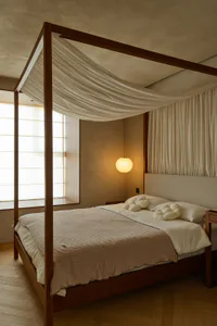 Modern minimalist bedroom with a large walnut four-poster bed, draped white fabric canopy, textured beige bedding, cloud-shaped pillows, and a glowing paper lantern pendant light beside a shoji-style window.