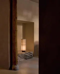 Warmly lit cylindrical table lamp placed on a grey marble ledge inside a recessed wall niche, beside a thick weathered wooden post with stone base in a minimalist interior.