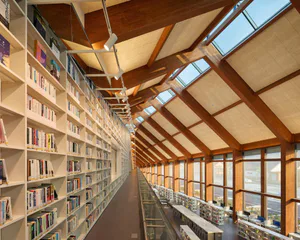 Long modern library corridor with tall white bookshelves on the left, wooden ceiling beams, large skylights, and reading tables on the right overlooking windows.