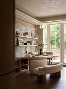 Elegant home office with a large curved mother-of-pearl desk, built-in shelving against a marble wall, cream leather bench, and large glass doors opening to a green balcony.