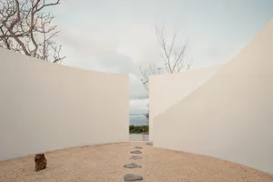 Minimalist outdoor courtyard with curved white concrete walls, gravel ground, irregular stone stepping path, a small dark rock sculpture, bare trees, and an opening revealing a sea view under a pale sky.