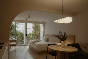Modern minimalist bedroom featuring a low platform bed with wooden headboard, round light oak dining table with black chairs, dark sofa, large arched doorway, and floor-to-ceiling glass doors opening to a balcony with greenery.