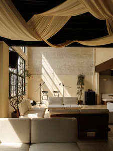 Modern industrial-style lounge with white modular sofas, draped fabric ceiling, exposed concrete walls, and dramatic natural light patterns.