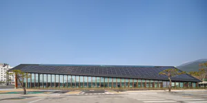 Exterior view of the modern Taichung Central Library with a large solar panel roof, extensive glass facade, and pine trees in the foreground under a clear blue sky.