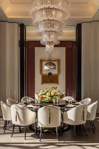 Elegant formal dining room featuring a large round black marble table surrounded by ten cream upholstered dining chairs with tassel details, a massive multi-tiered crystal chandelier, and a large floral centerpiece.