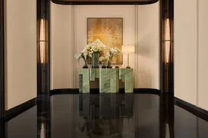 Modern luxury entryway featuring a green marble console table with white flower arrangements, large abstract gold painting, and dramatic vertical wall sconces reflected in a glossy black floor.