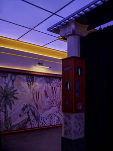 Interior view of a surreal art space featuring a large illustrated mural of a spotted horse and fantastical plants, paired with a multi-material column that combines a classical capital, red wood, marble, and decorative inlays under purple ceiling lighting.