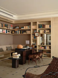 A warm and elegant home library featuring floor-to-ceiling built-in bookshelves, a large walnut desk, two mid-century wooden armchairs, a built-in window seat with checkered cushions, and a brown leather sling chair in the foreground.