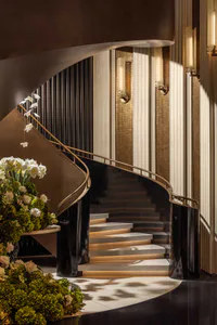 Elegant curved grand staircase with white marble steps featuring integrated lighting, polished dark balustrades, brass handrails, and large floral arrangements of white orchids and green hydrangeas in a luxurious hotel lobby.