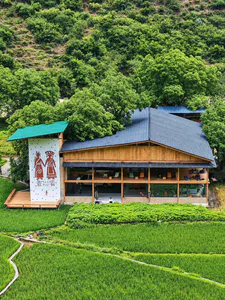 A large two-story wooden building with a dark slate roof, open ground floor, and a white mural wall featuring red traditional figures, surrounded by green rice paddies and forested mountains.