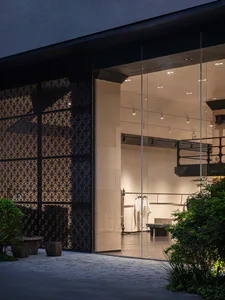 Modern boutique storefront at dusk with large glass windows, a black perforated metal screen wall, stone pavers, and green landscaping.